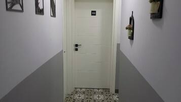 Un couloir étroit avec des carreaux de sol à motifs, une porte blanche portant un numéro, et des décorations murales, notamment des œuvres d’art encadrées et une petite étagère avec des fleurs.