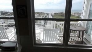 Ein Zimmer mit großem Fenster, das auf eine Terrasse mit Gartenmöbeln und Blick auf das Meer hinausgeht.