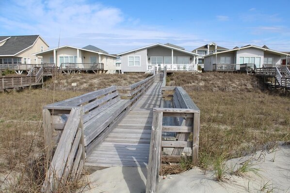 A wooden boardwalk leading to beach houses with a sandy beach and dunes in the background.