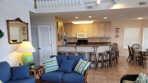 A living room with a blue sofa, a coffee table, and a dining area with bar stools and a table. - Spacious Oceanfront Retreat -Steps to Sand & Pool Access! By Williamson Realty (Ocean Isle Beach)