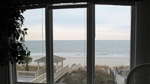 A room with a large window overlooking a beach, a chair, and a potted plant. - Spacious Oceanfront Retreat -Steps to Sand & Pool Access! By Williamson Realty (Ocean Isle Beach)