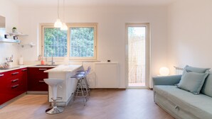 A modern kitchen with red cabinets, a white countertop, and a bar stool. There is a window with a view of trees, and a light-colored sofa in the living area. - MONDELLO COTTAGE ULIVO 1 | Beach Apartment with Private Garden & Parking (Palermo)