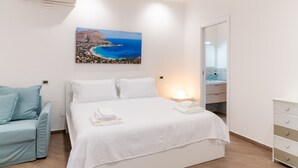 A hotel room with a bed, a seating area, a painting of a coastal landscape, and a bathroom with a mirror and sink. - MONDELLO COTTAGE ULIVO 1 | Beach Apartment with Private Garden & Parking (Palermo)
