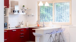 A modern kitchen with red cabinets, white countertops, and a window with a view of greenery. - MONDELLO COTTAGE ULIVO 1 | Beach Apartment with Private Garden & Parking (Palermo)