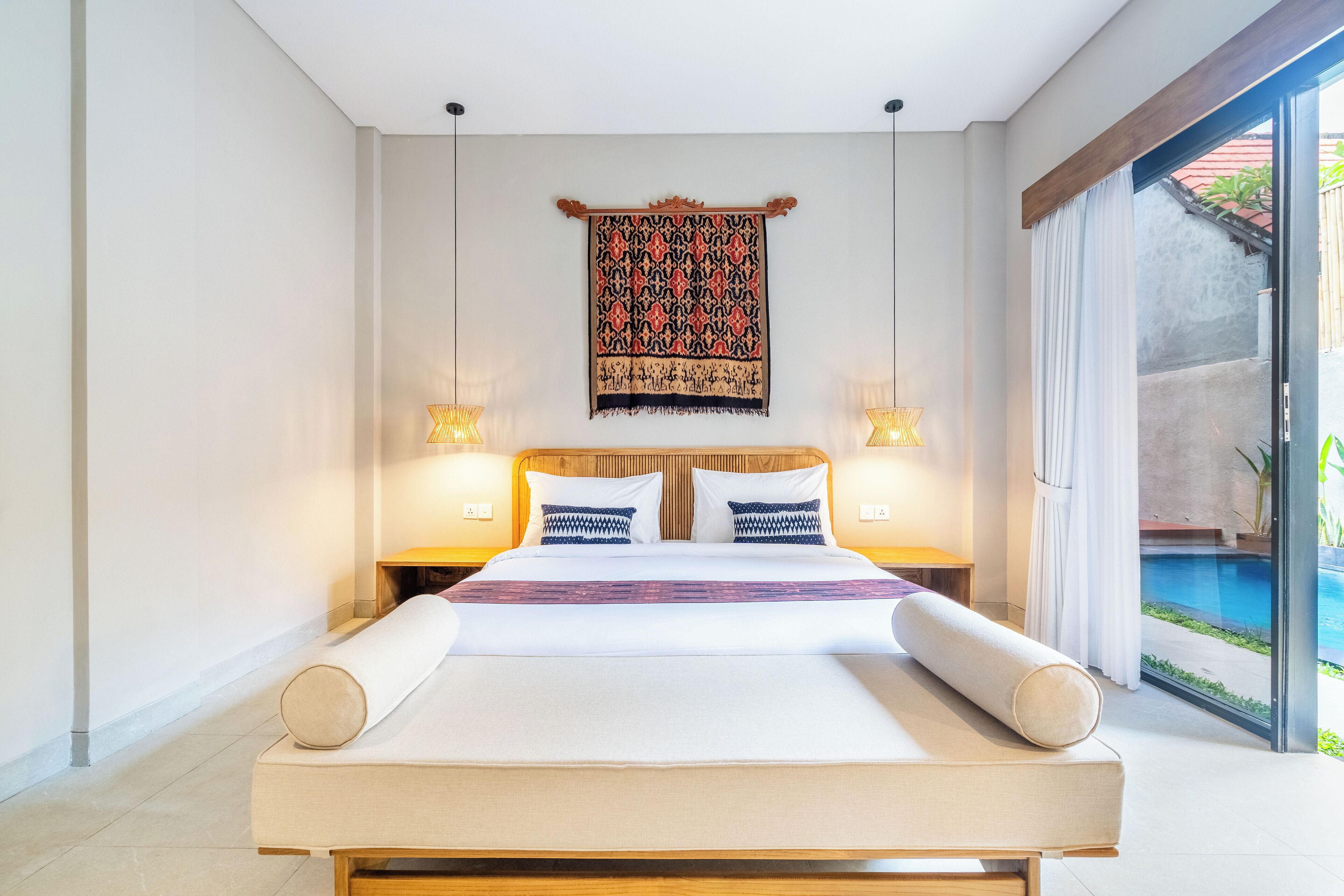 A modern bedroom with a large bed, two bedside tables with lamps, a patterned tapestry, and a view of a swimming pool.