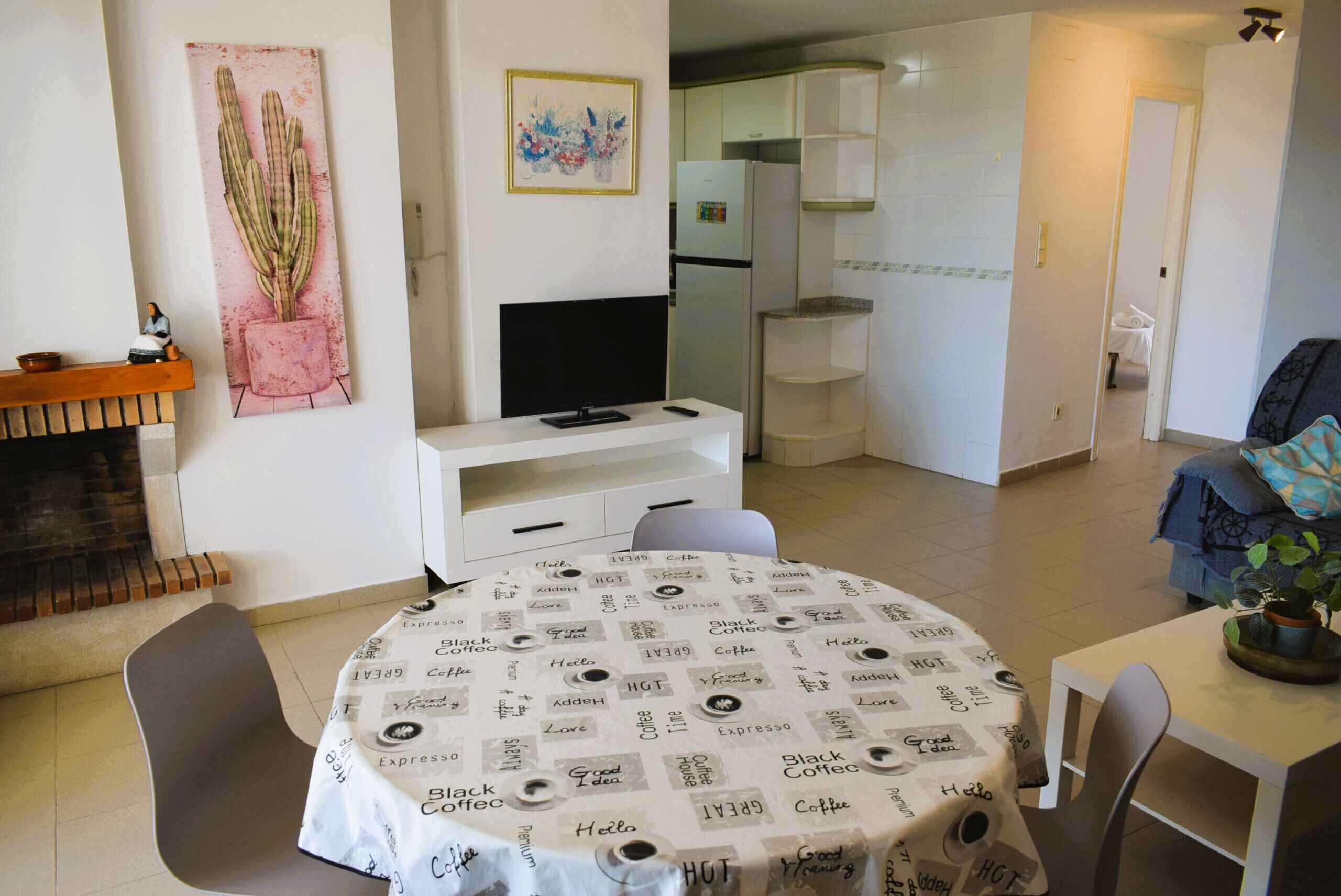 A dining area with a round table covered in a coffee-themed tablecloth, a television on a white cabinet, and a cactus painting on the wall.