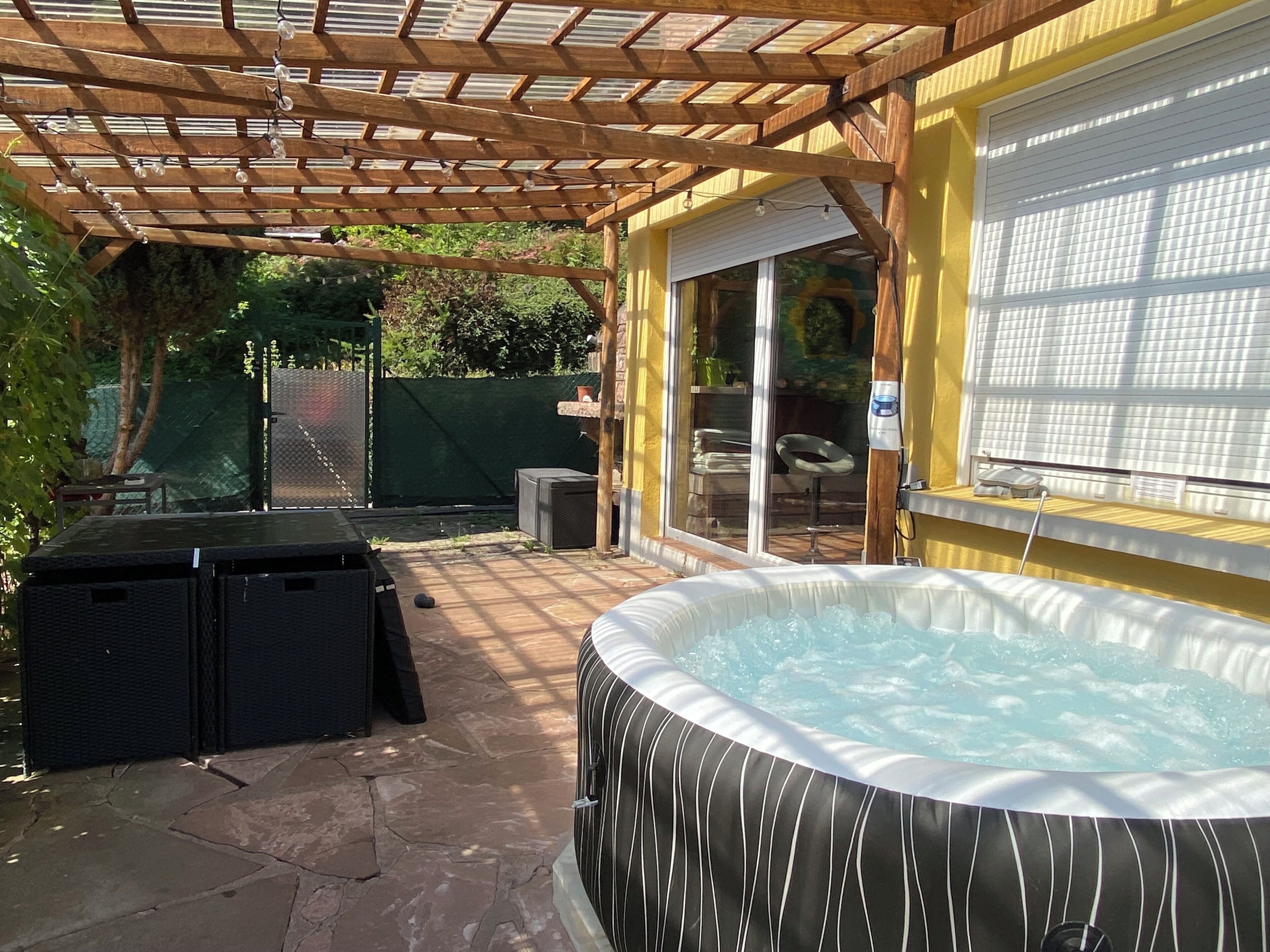 Un jacuzzi bajo una pérgola de madera, una zona de patio con suelo de piedra y un espacio de estar cubierto con una mesa y sillas.