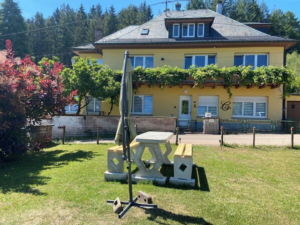 A yellow building with a sloped roof, a door, and windows. There is a bench and a table in the foreground.