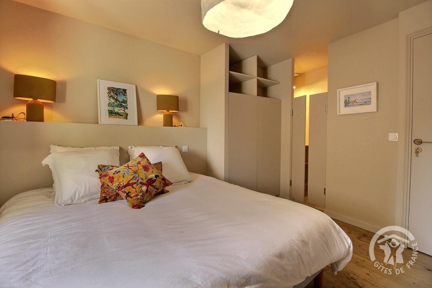 A bedroom with a bed, two bedside lamps, a wall-mounted shelf with a picture, and a wardrobe with a mirror.