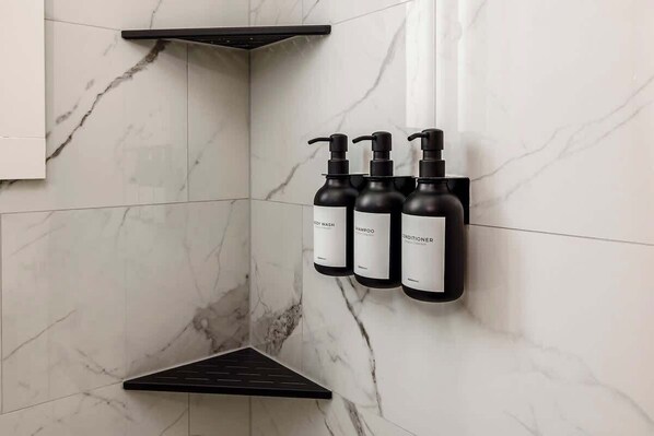 A bathroom with marble walls, a black shelf, and three black bottles labeled for hot wash, shampoo, and conditioner.