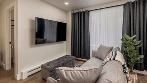 Een moderne woonkamer met een flatscreen televisie aan de muur, een grijze bank, een bijpassend voetenbankje en een potplant.
