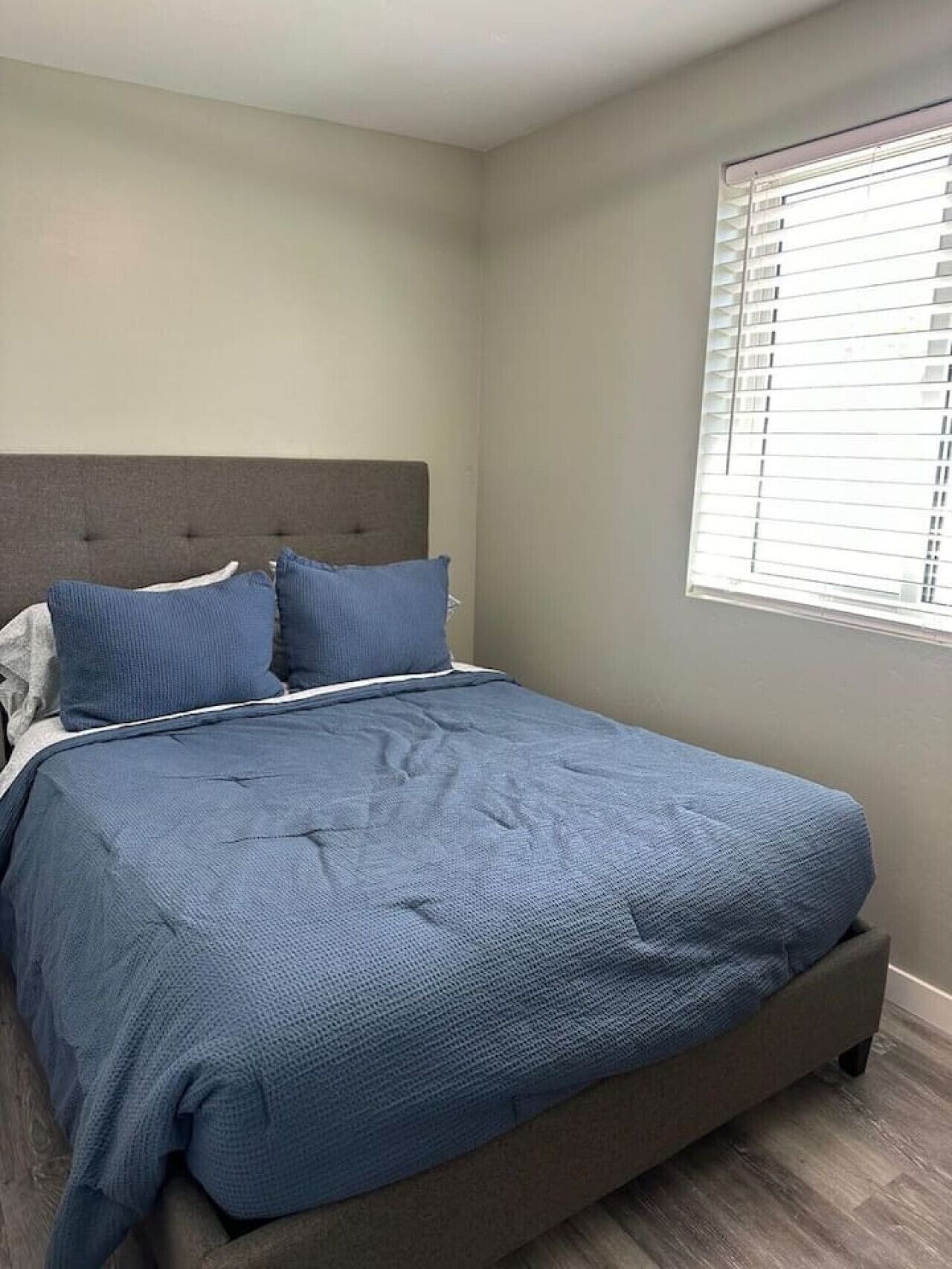 A bedroom with a bed, grey headboard, and a window with blinds.