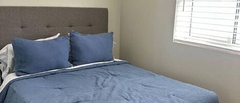 A bedroom with a bed, grey headboard, and a window with blinds.