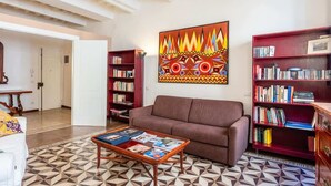 Ein Wohnzimmer mit einem braunen Sofa, einem Couchtisch mit Zeitschriften, einem Bücherregal und einem farbenfrohen, abstrakten Gemälde an der Wand.