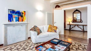 Ein Wohnzimmer mit weißem Sofa, bunten Kissen, einem Couchtisch mit Zeitschriften, einem geometrisch gemusterten Boden, einem großen abstrakten Gemälde und einem Spiegel mit Holzrahmen.