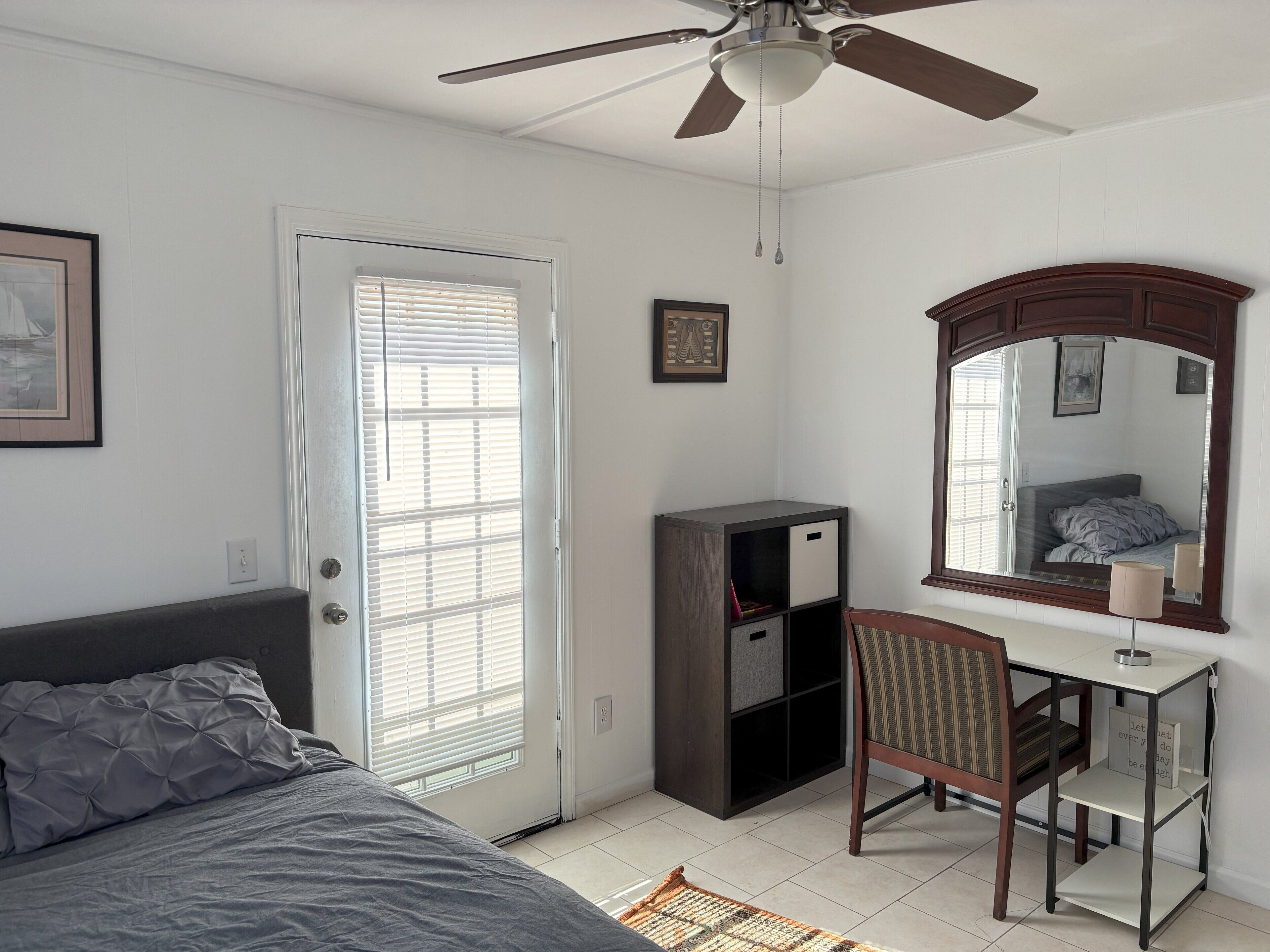 A bedroom with a bed, a desk with a chair, a wardrobe, a ceiling fan, and a large mirror.