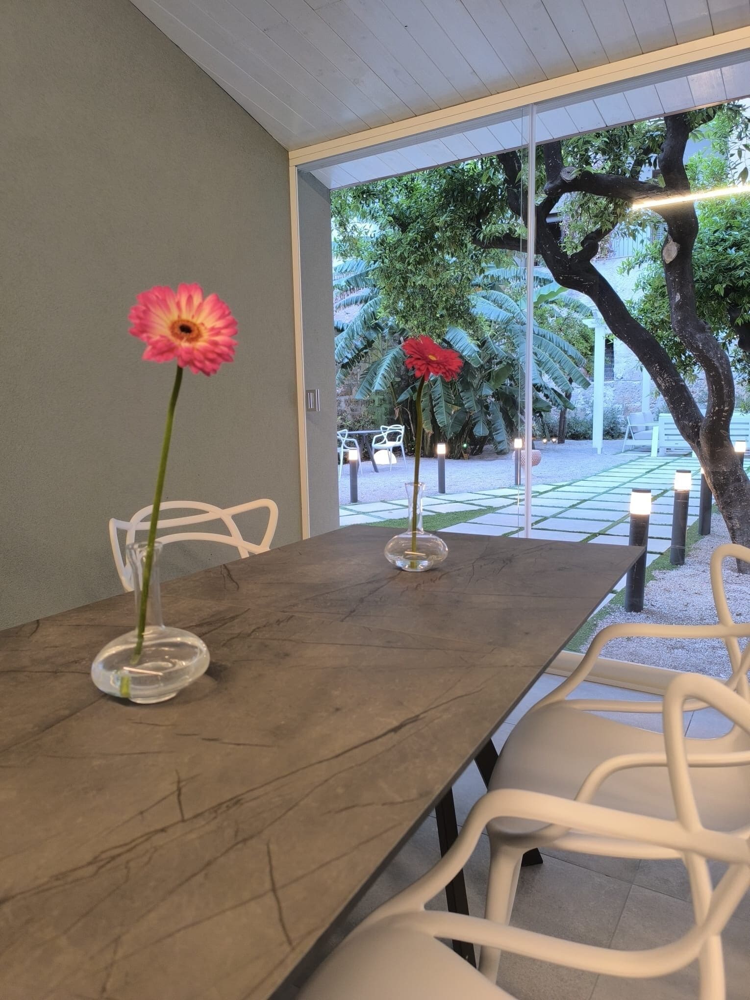 Un coin repas avec une table en bois et des chaises blanches, une fleur rose dans un vase transparent, et une vue sur un jardin avec des arbres et des chaises à l’extérieur.