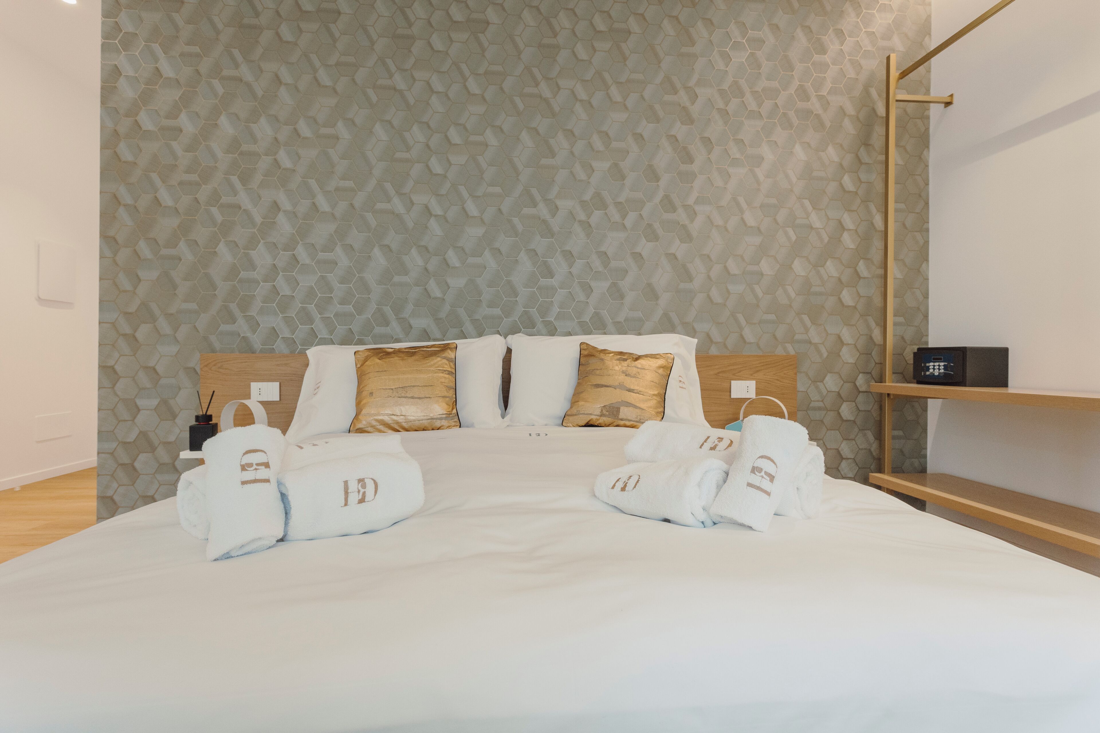 A hotel room with a bed, two pillows, a wooden headboard, a bedside table with a black object, and a wall with a honeycomb pattern.