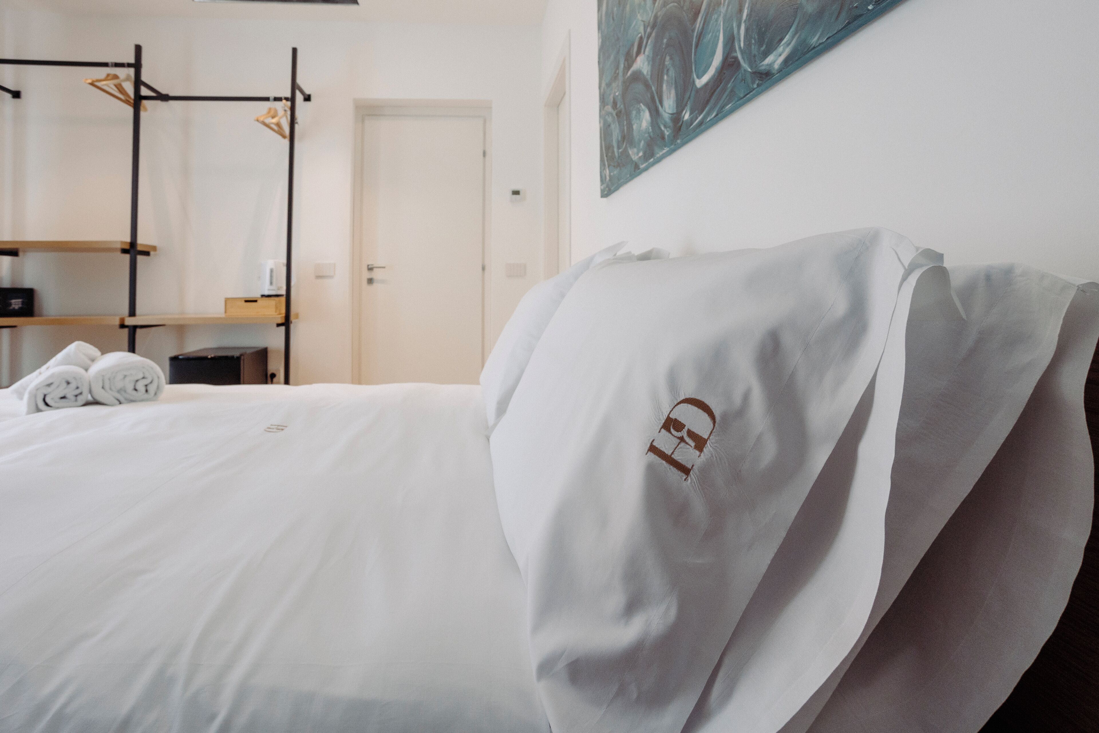 Ein ordentlich bezogenes Bett mit weißen Laken und einem Kopfkissen mit Logo, eine Holzregaleinheit und eine Tür in einem Hotelzimmer.