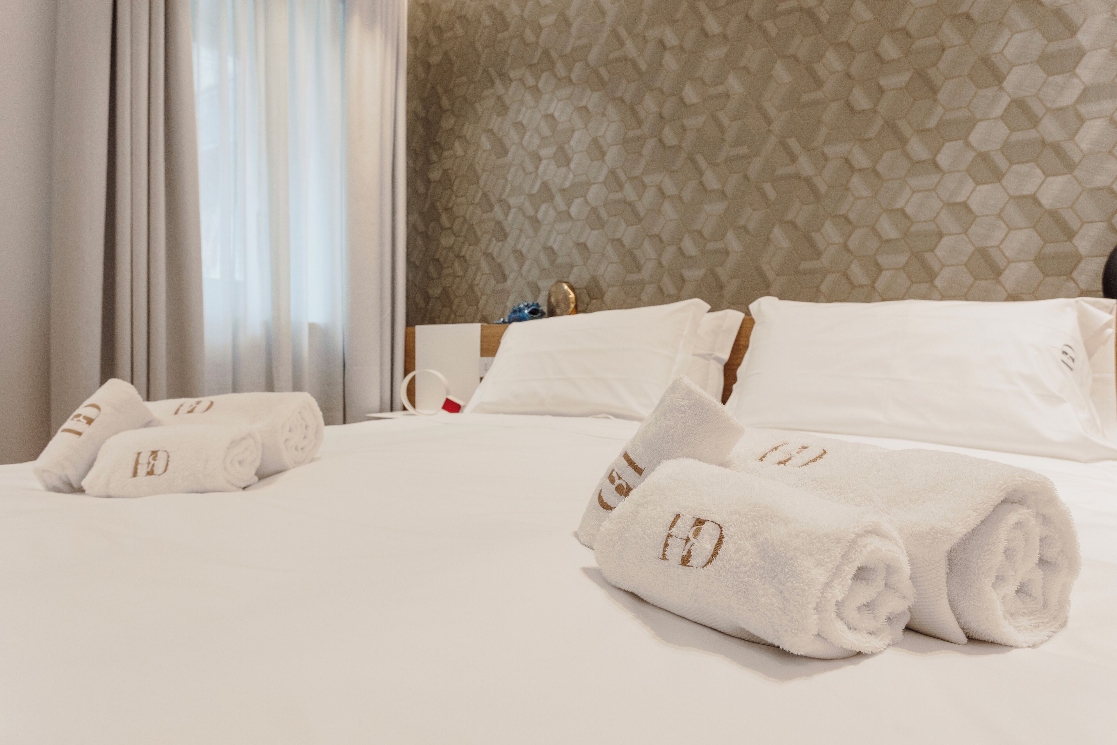 A hotel room with a neatly made bed, rolled white towels with a logo, and a textured hexagonal wall.
