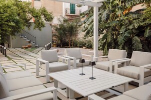 Un patio con mobiliario blanco, incluyendo sofás y una mesa, rodeado de vegetación y una escalera.