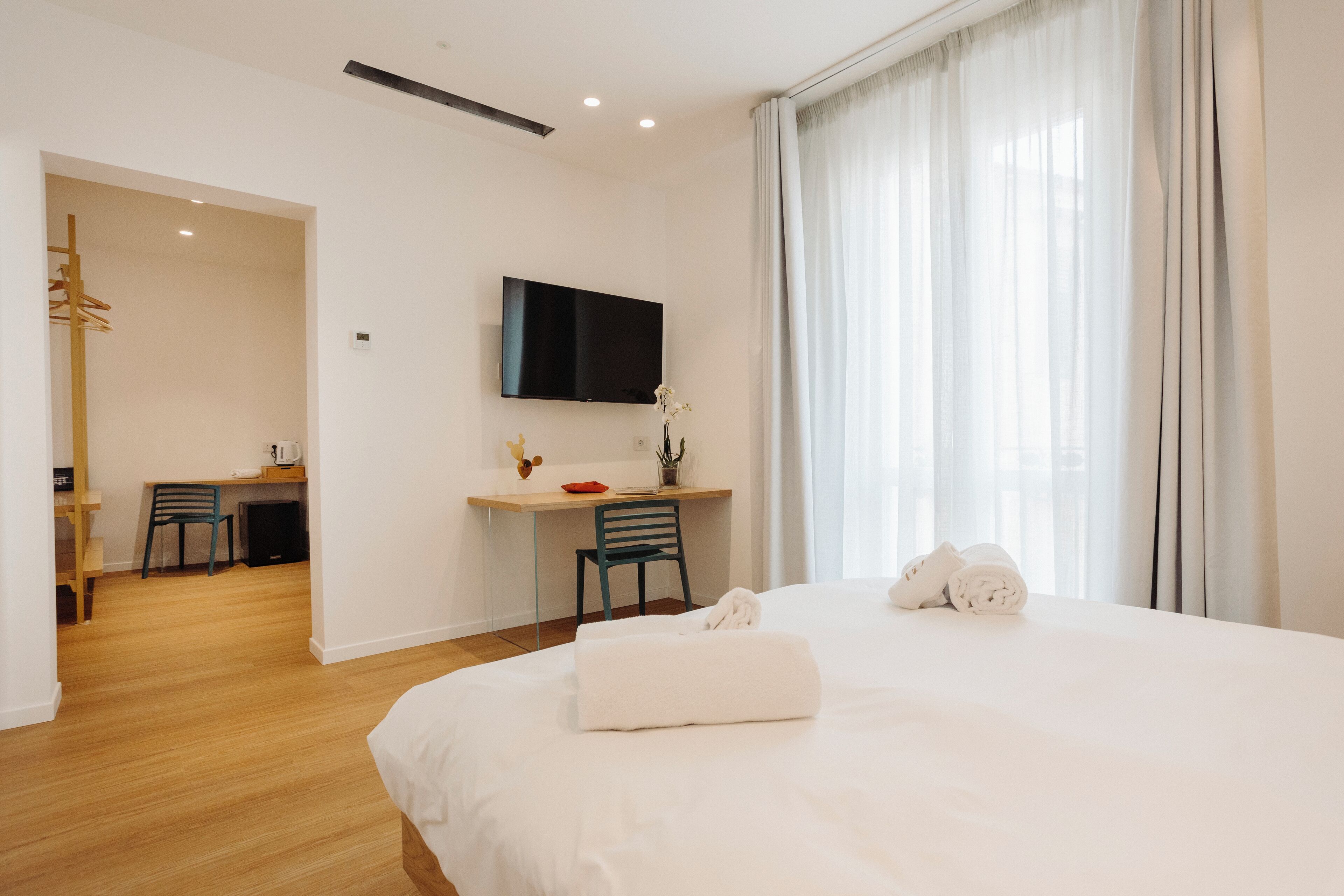 Habitación de hotel con cama, escritorio con silla, televisor montado en la pared y ventana con cortinas.