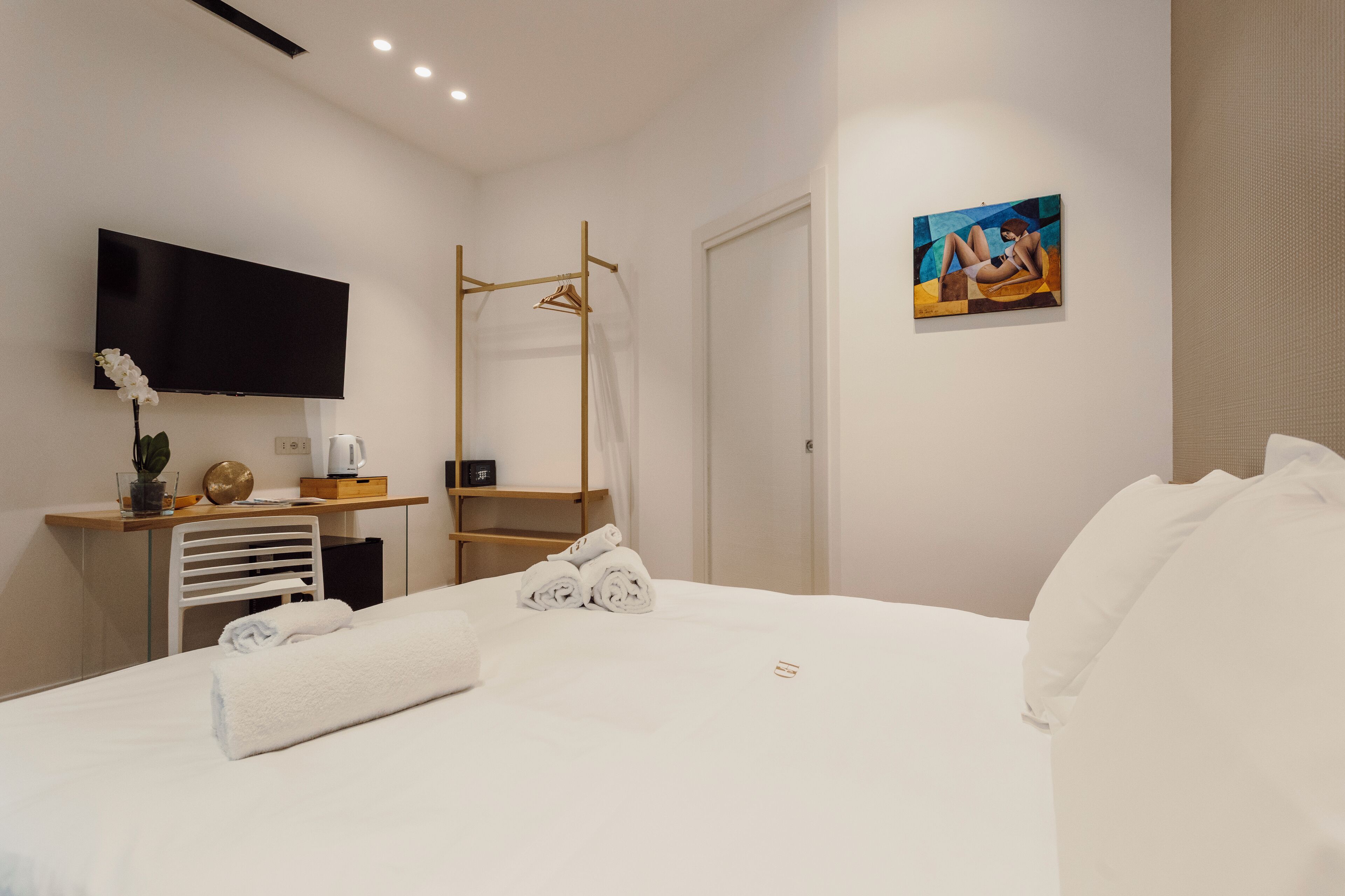 A hotel room with a bed, a TV, a desk with a chair, a painting on the wall, and a shower area.