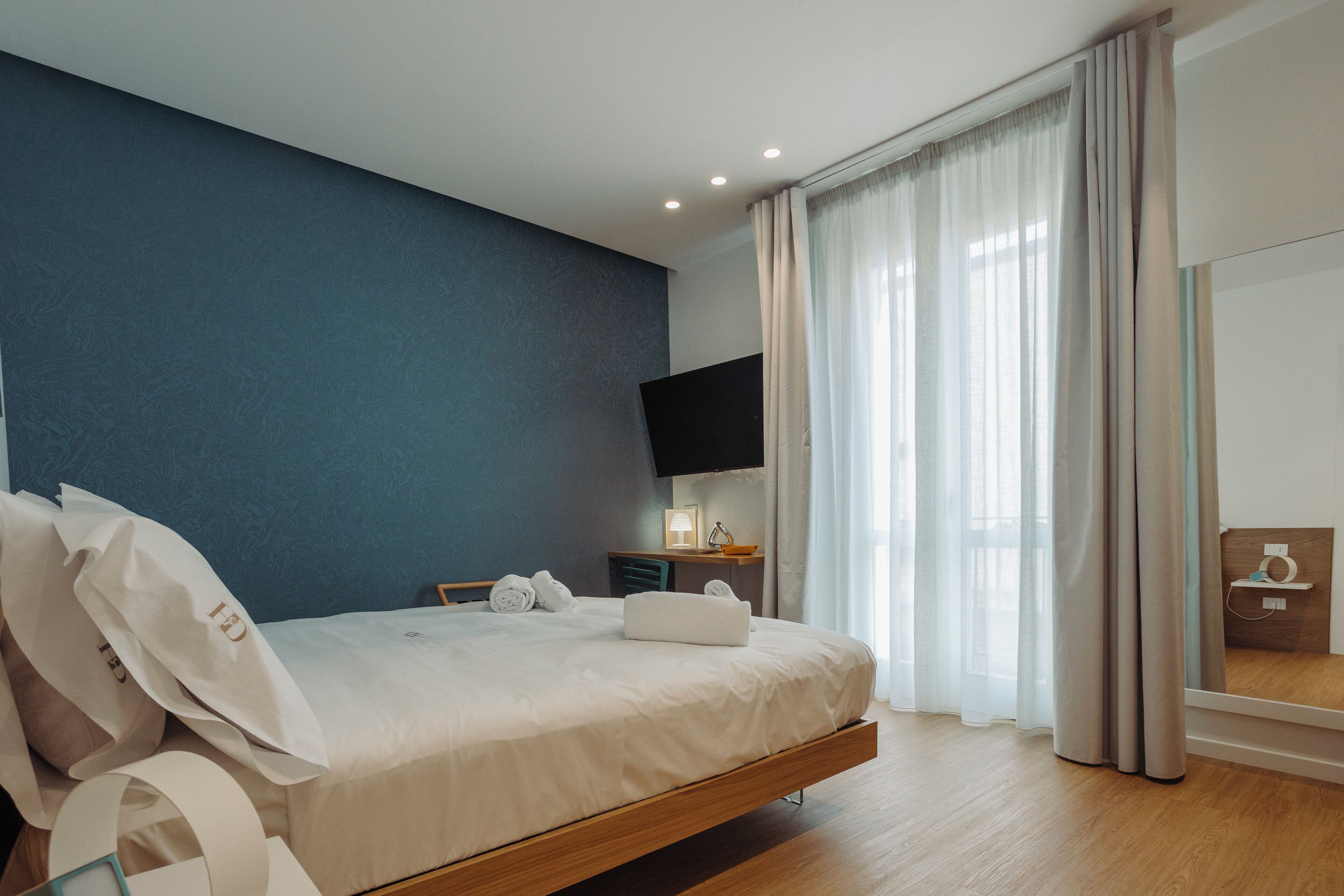 A modern hotel room with a large bed, a desk, a flat-screen TV, and a large window with curtains.