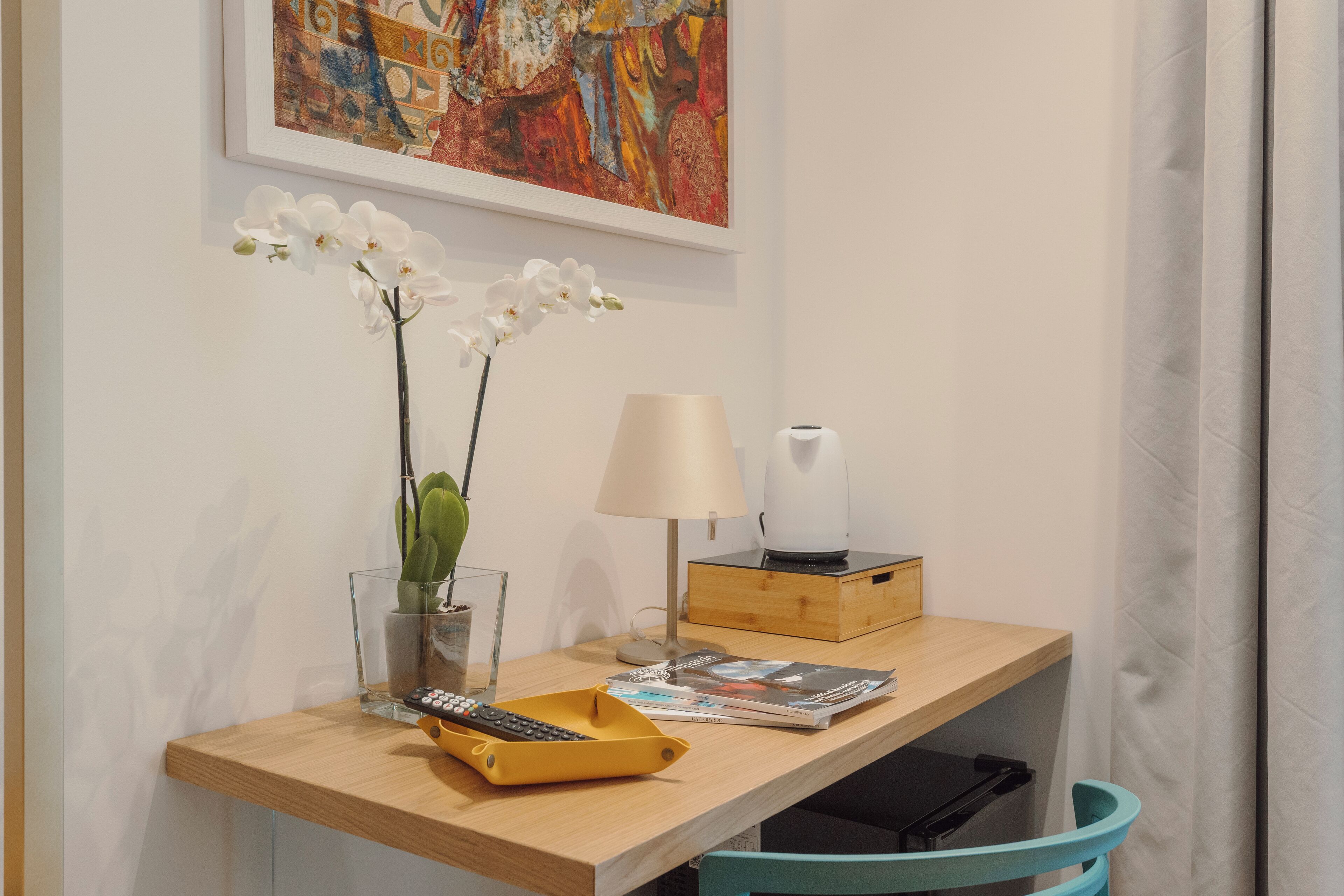 A wooden desk with a remote control, a lamp, a magazine, and a vase with white orchids.