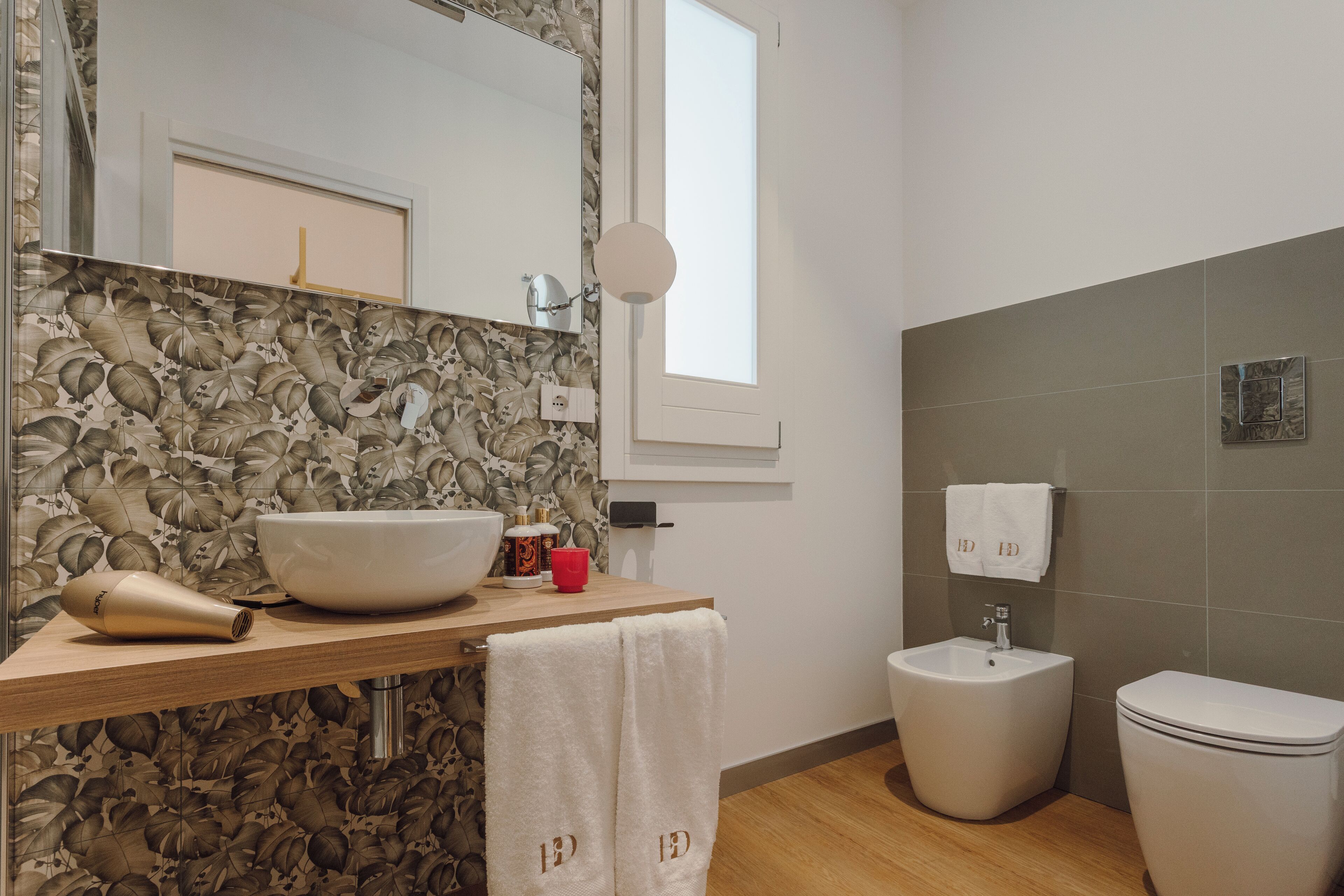 Een badkamer met een grote spiegel, een wastafel, een bidet en een toilet. Een handdoek hangt aan de muur en er is een raam boven de wastafel.