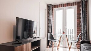 En moderne stue med et fladskærms-TV på et træskab, et glasbord med to stole, og en balkon med gelænder af glas.