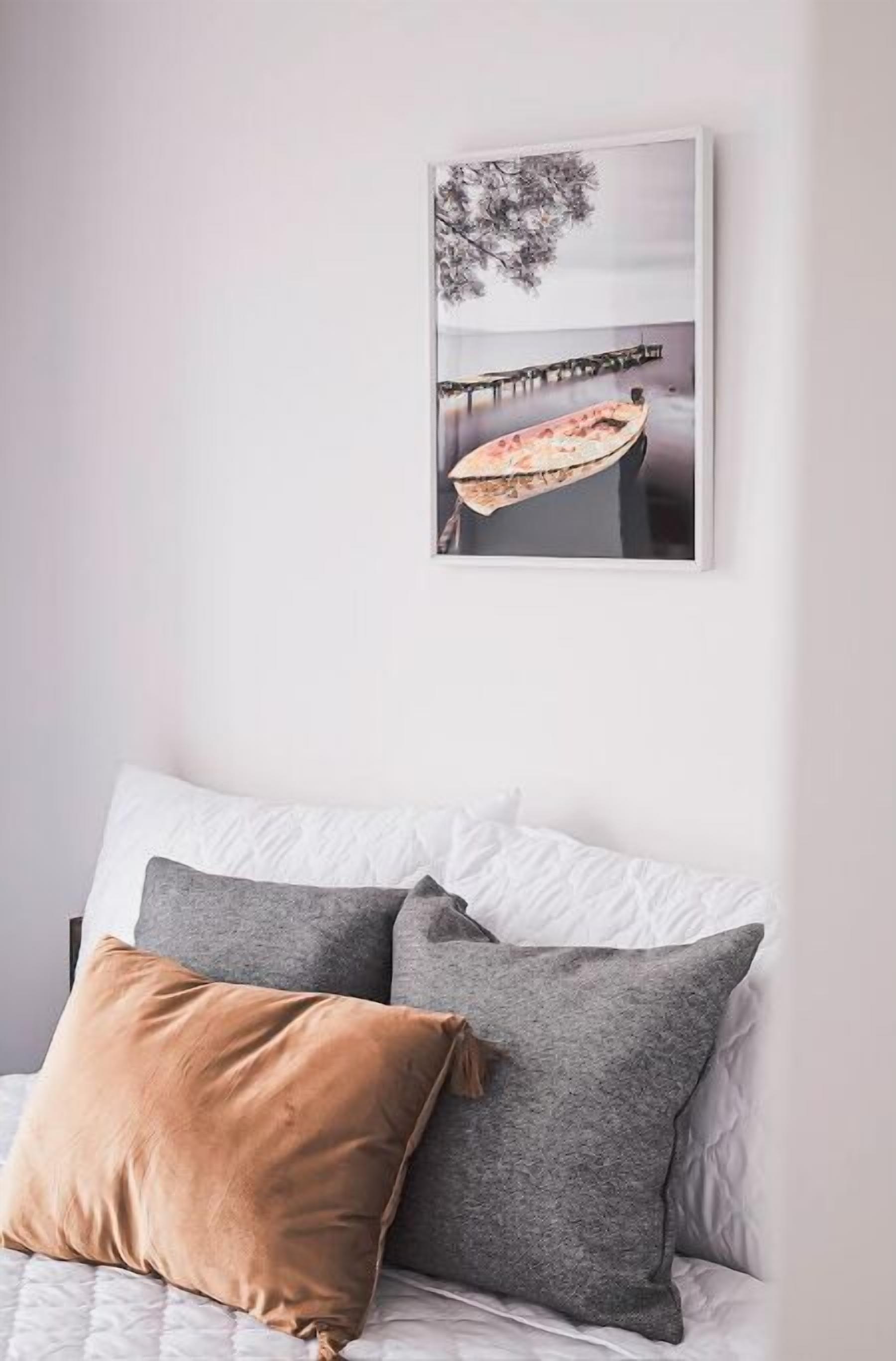A bed with white and grey bedding, a brown pillow, and a framed picture of a boat on a pier.