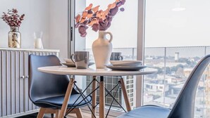 Un espace repas moderne avec une table ronde, deux chaises, un vase de fleurs et une vue sur la ville grâce à de grandes fenêtres.