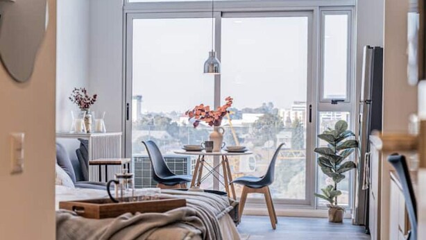 Une chambre moderne dotée d’une grande porte-fenêtre coulissante donnant sur un balcon, d’un lit avec une tête de lit en bois, d’une petite table avec un vase de fleurs et d’une plante en pot sur le balcon.