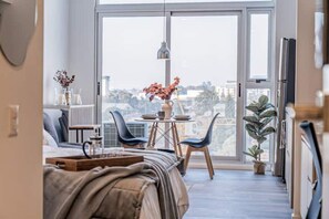 A modern bedroom with a large sliding glass door leading to a balcony, a bed with a wooden headboard, a small table with a vase of flowers, and a potted plant on the balcony.