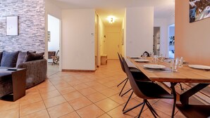 A modern dining area with a long wooden table set for six, flanked by black chairs. A stone-patterned wall and a gray sofa with cushions are visible in the background. - Cassio (Annecy)