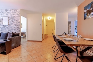 Un espace de salle à manger moderne, avec une longue table en bois dressée pour six personnes, entourée de chaises noires. On aperçoit, à l’arrière-plan, un mur imitant la pierre et un canapé gris agrémenté de coussins.