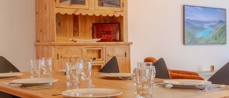 Un coin repas avec une table en bois dressée pour quatre personnes, des chaises noires, un buffet en bois doté de portes en verre et un tableau représentant un paysage, accroché au mur.