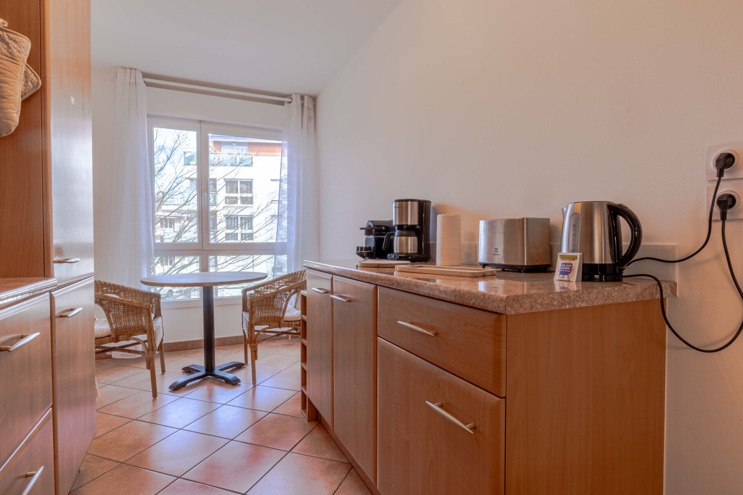A compact kitchen with a dining area, featuring a coffee maker, kettle, and a small table with chairs.