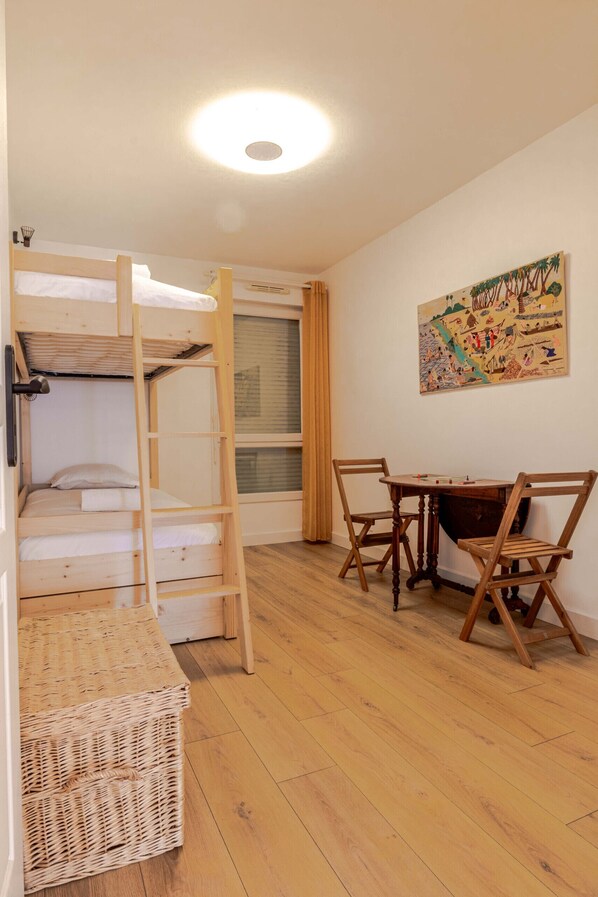 Une pièce avec un lit superposé, une table en bois avec des chaises, un panier en osier et un mur orné d’un tableau coloré.