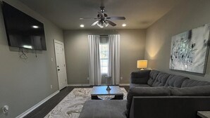 A living room with a dark wood floor, a grey sectional sofa, a coffee table, a ceiling fan, a flat-screen TV mounted on the wall, and a window with curtains. - Charming 1-bedroom apartment with WiFi, AC in gorgeous Sour Lake #211 (Sour Lake)