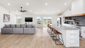 A modern kitchen with a central island, bar stools, and a dining area with a grey sofa. - Bungalow on 3 Acres - Hot Tub& Firepit (McKinney)
