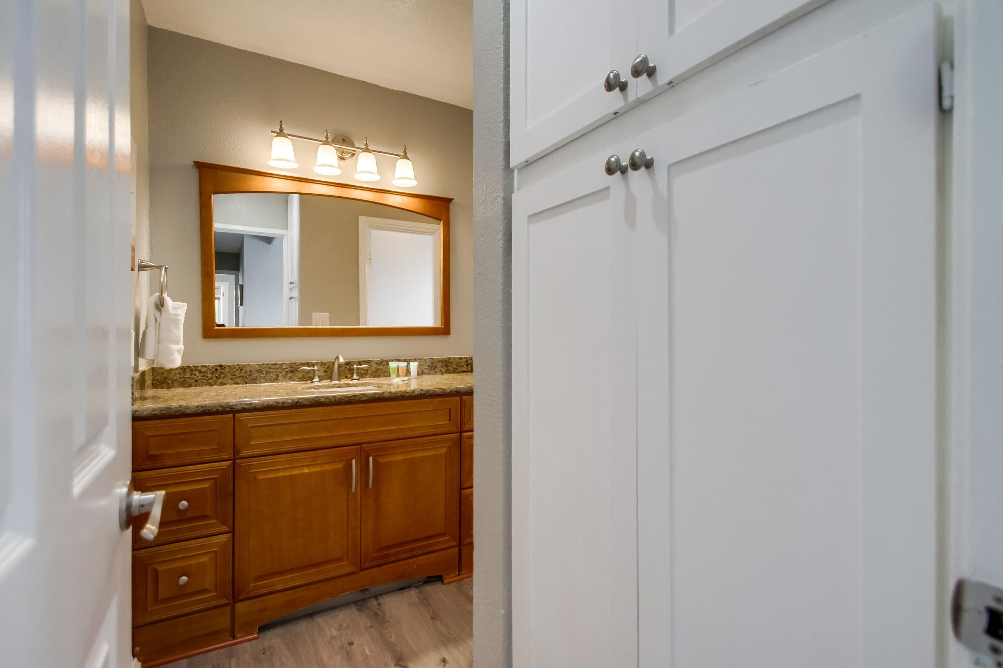 A bathroom with wooden cabinets, a granite countertop, a large mirror with lights, and a white door.
