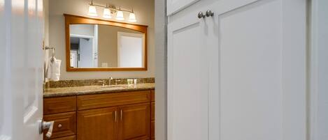 A bathroom with wooden cabinets, a granite countertop, a large mirror with lights, and a white door.