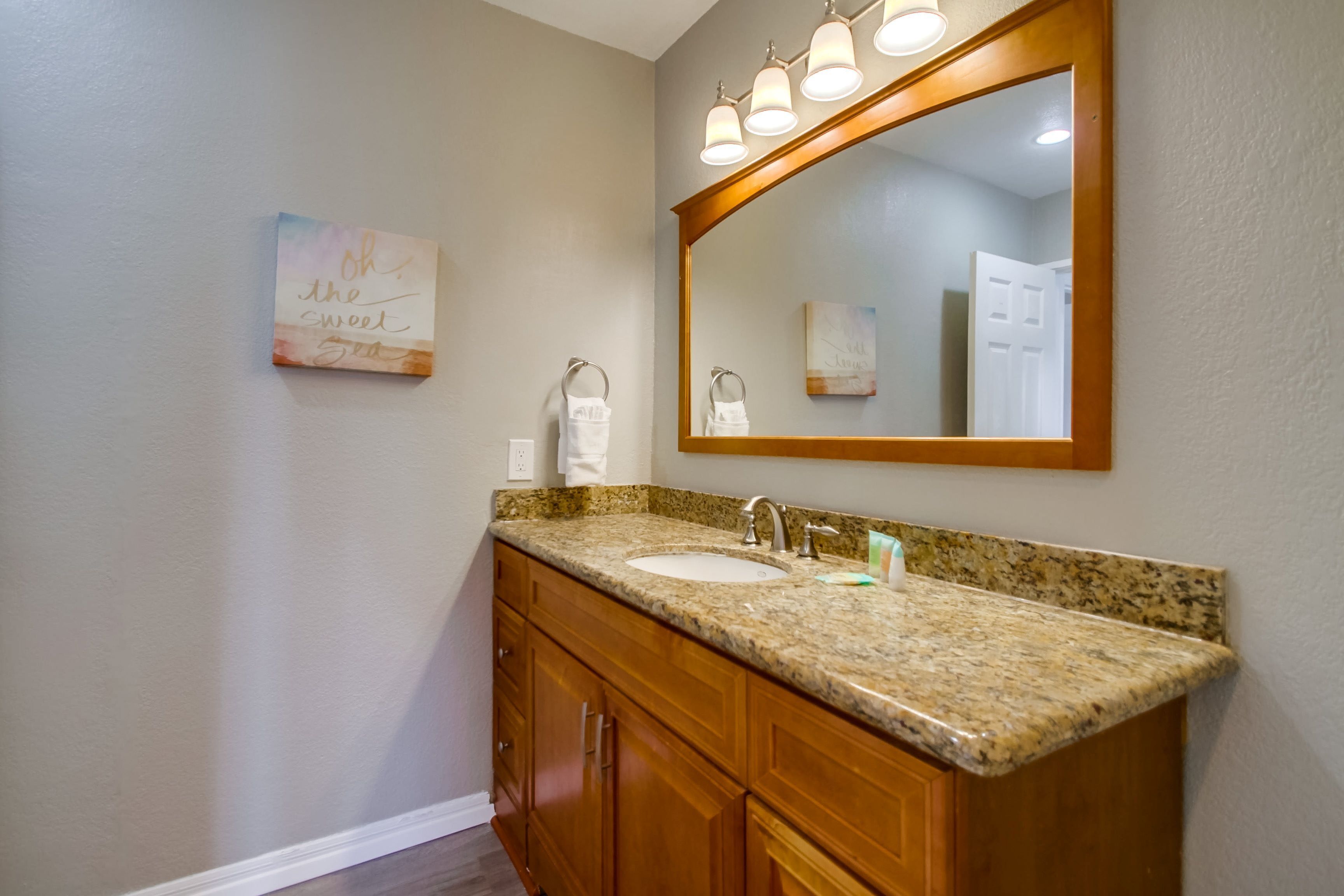A bathroom with a granite countertop, two sinks, wooden cabinets, and a large mirror with lights above.