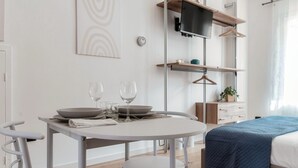 A modern room with a round dining table, two chairs, a bed with a blue cover, a TV on a wooden shelf, and a wall-mounted light switch.