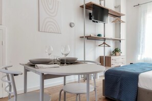 A modern room with a round dining table, two chairs, a bed with a blue cover, a TV on a wooden shelf, and a wall-mounted light switch.