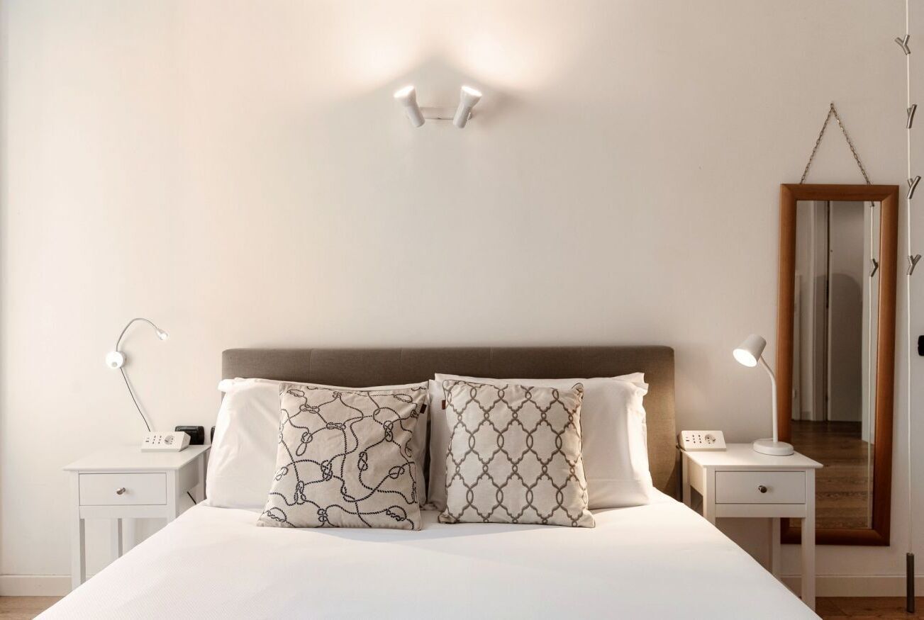 A neatly arranged bedroom with a bed, two bedside tables, a nightstand, a wall-mounted light, and a mirror.