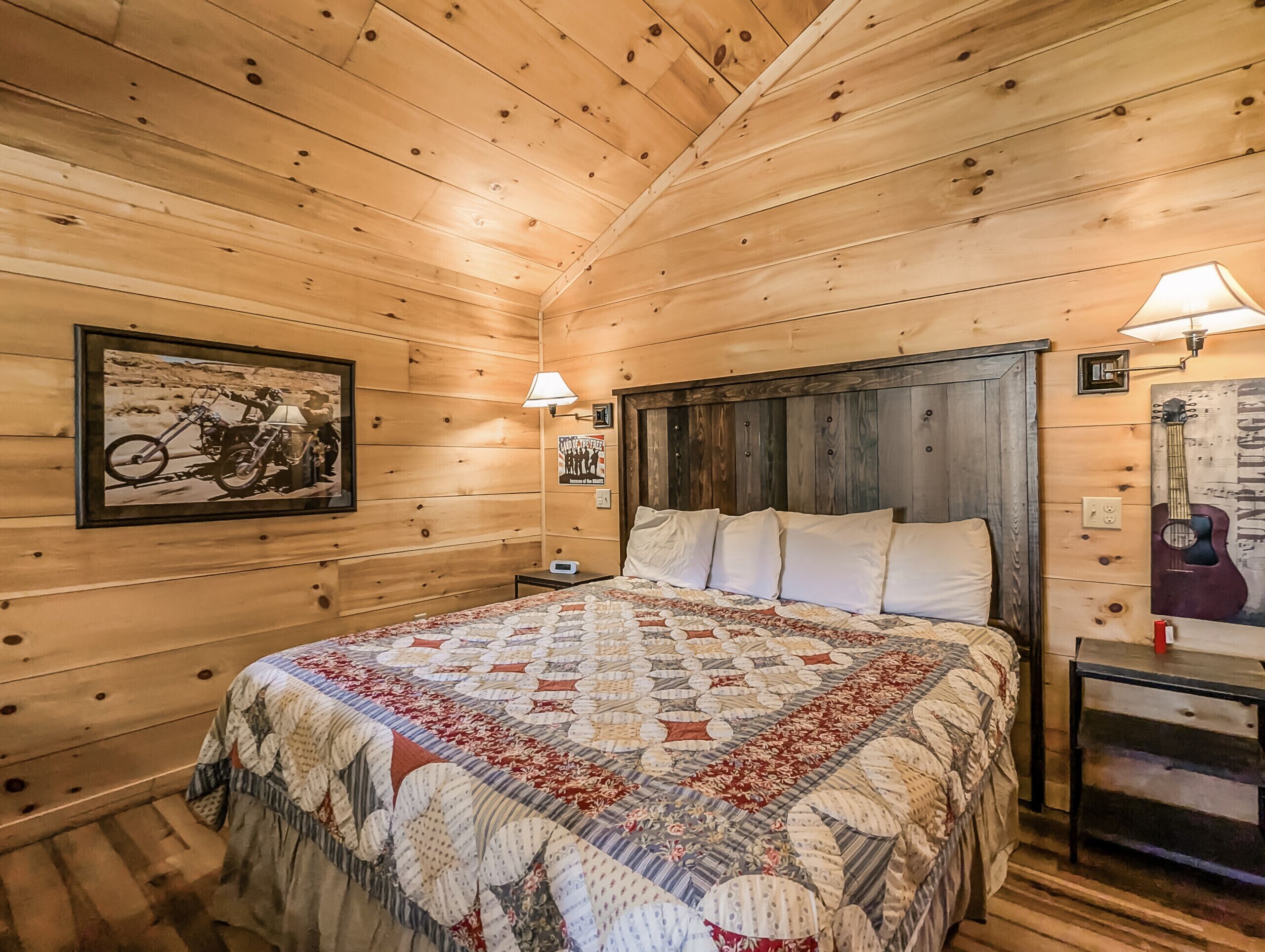 A cozy wooden cabin bedroom with a large bed, a framed motorcycle picture, a guitar, and a quilted bedspread.