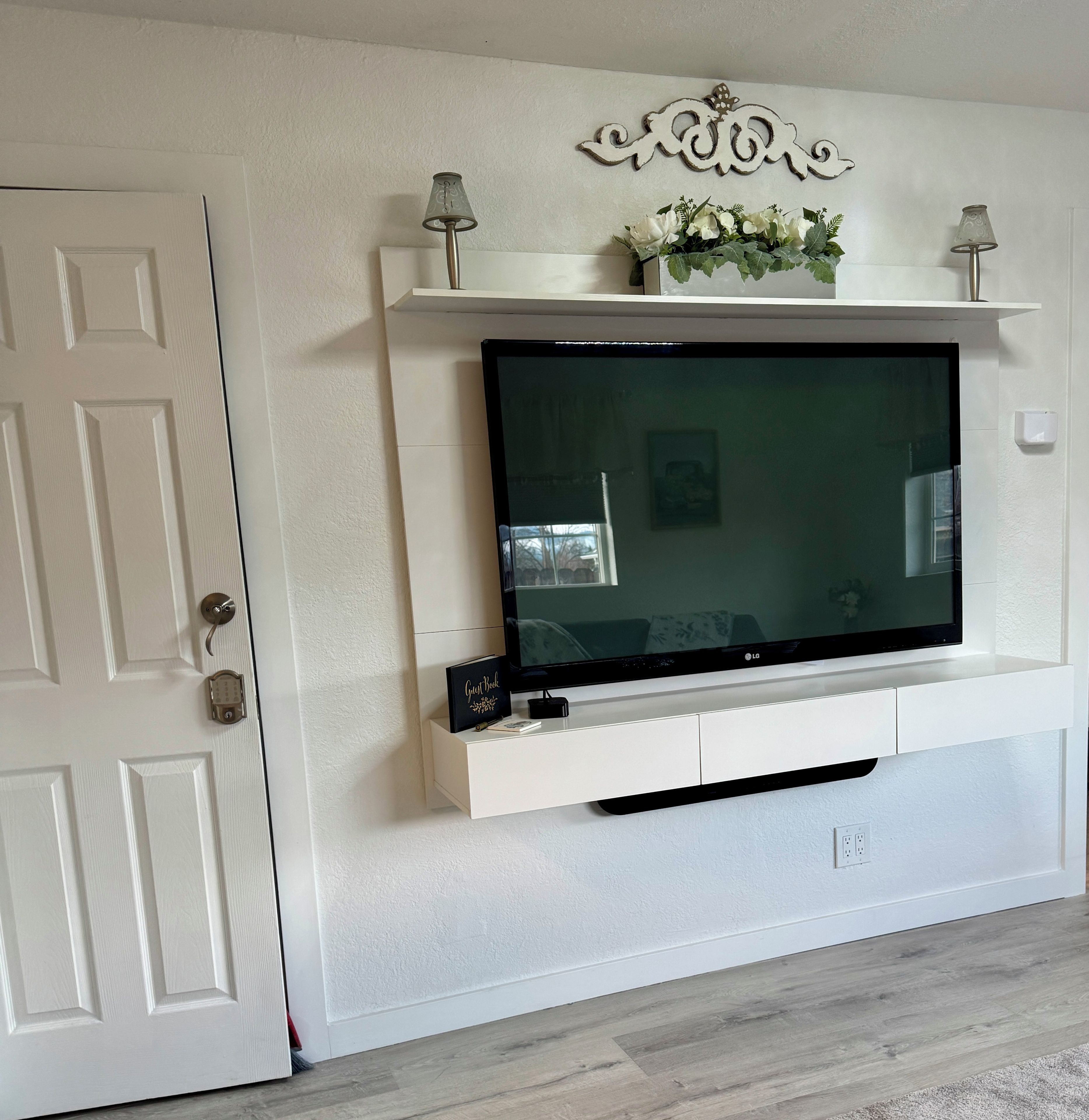 Een woonkamer met een televisie aan de muur, een witte deur en een decoratief kunstwerk boven de televisie.